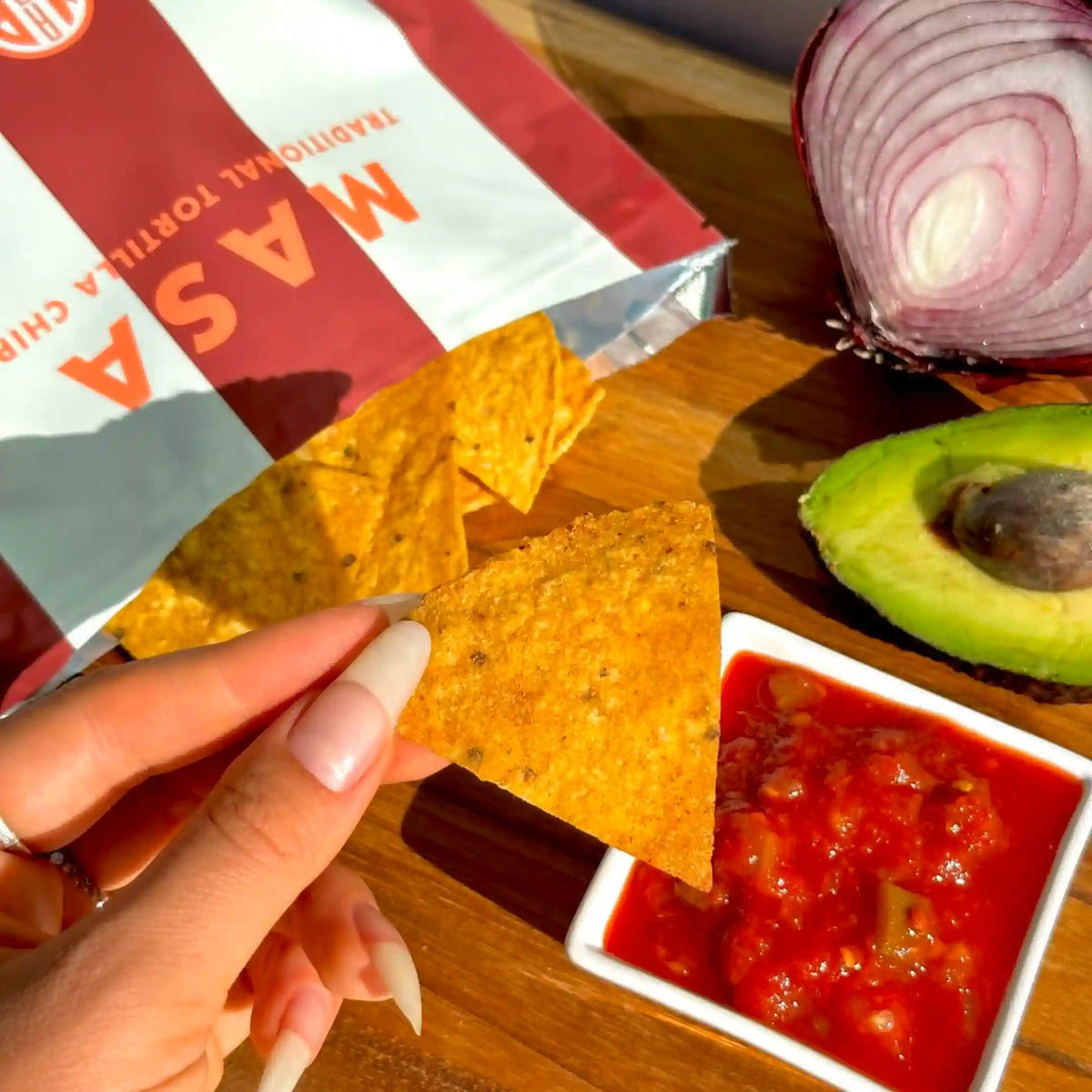 A MASA chip scooping salsa, with guacamole and an open bag of MASA Original